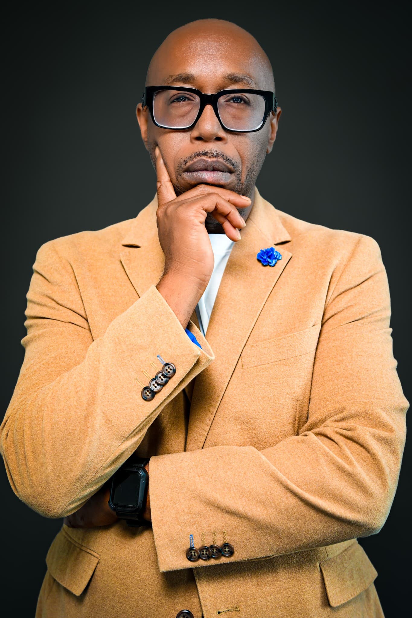 Bald Black man in a tan blazer and glasses, posing thoughtfully with hand on chin.